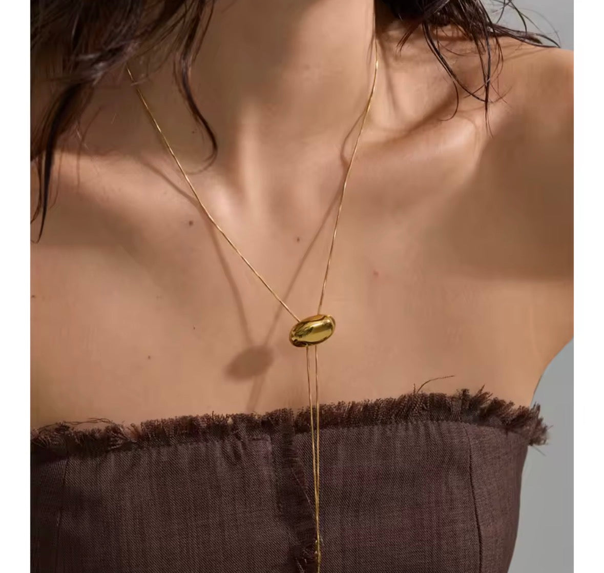 A close-up of a model wearing a delicate gold chain necklace with a small, polished gold oval pendant and a long drop chain extending down to a dark brown fringed strap.