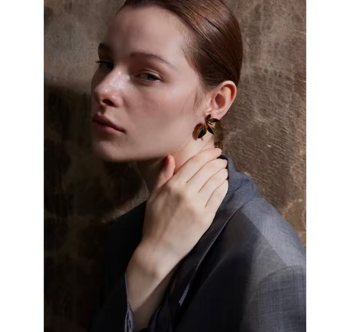 A close-up portrait of a woman wearing a large, organic-shaped gold earring with dark brown stone inlay, set against a textured brown background.