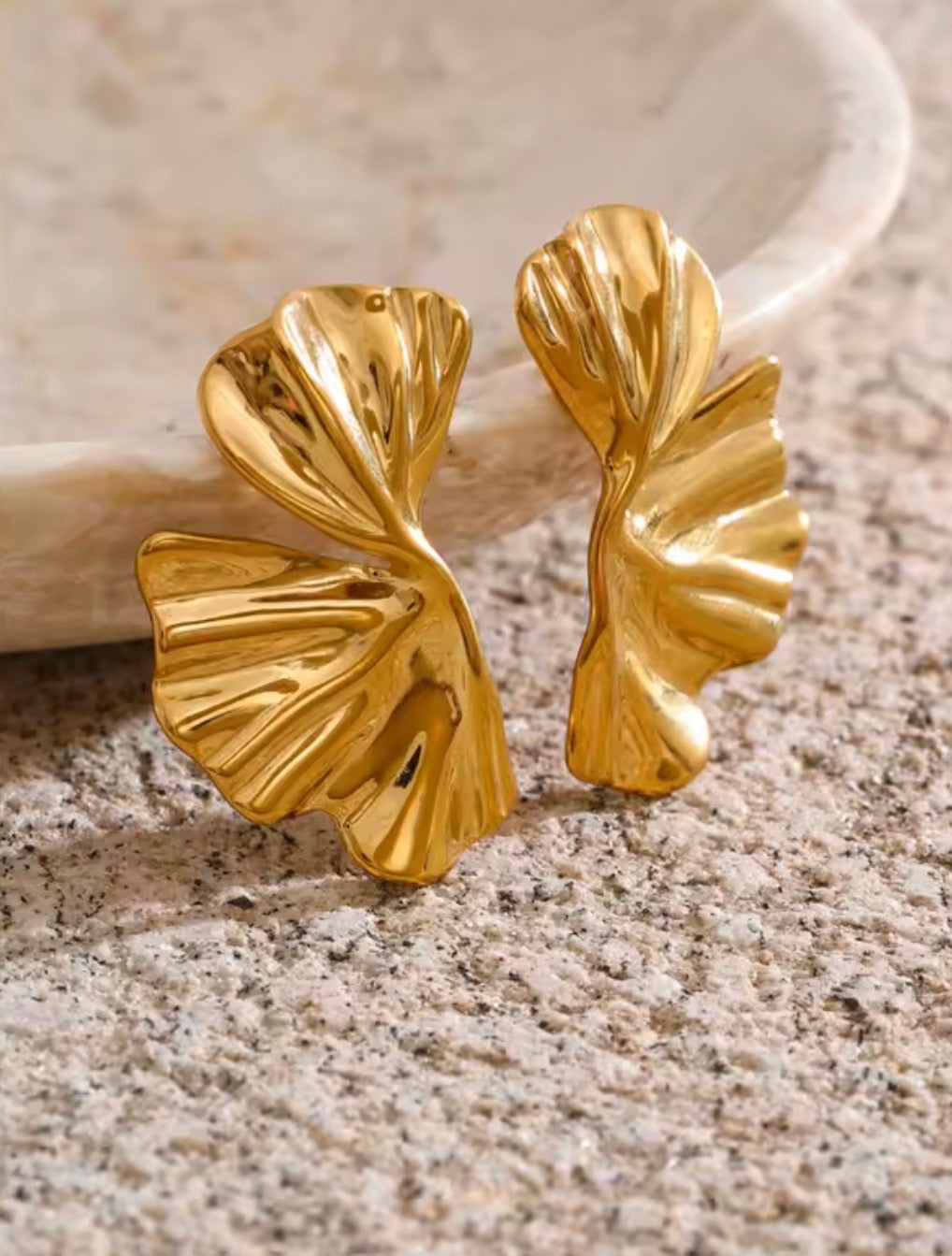 A pair of gold leaf-shaped stud earrings with a textured, ribbed surface, displayed on a speckled stone surface next to a light-colored ceramic dish.
