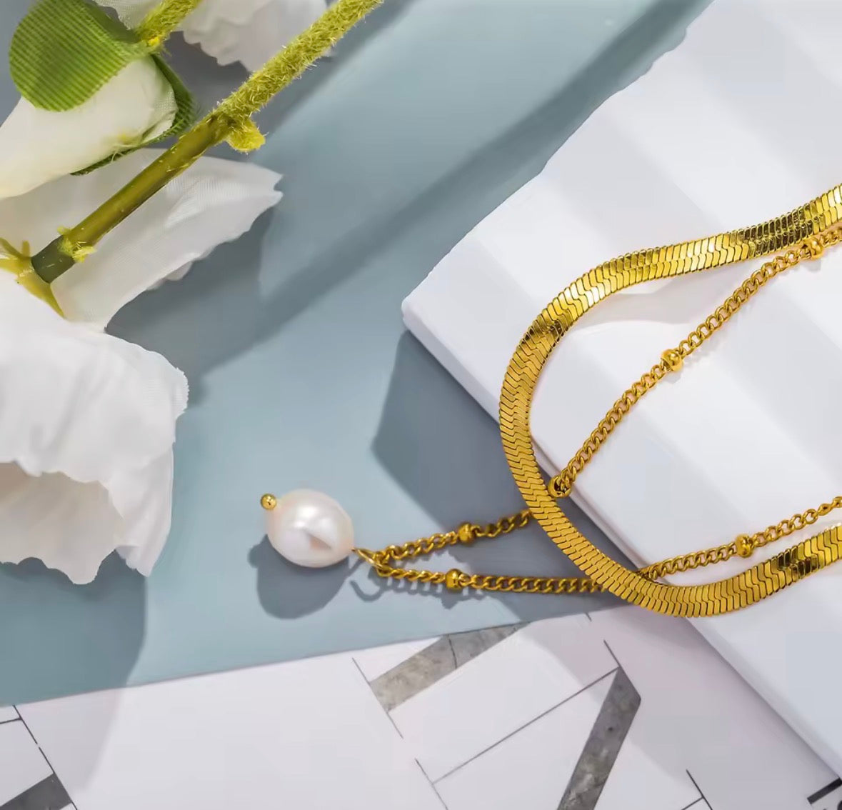 Gold chain necklace with a pearl pendant on a light blue surface with white flowers.