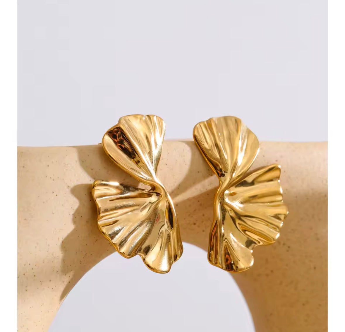 A pair of gold-toned, textured leaf-shaped earrings displayed on a beige jewelry stand against a light gray background.