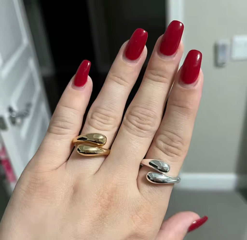 Hand with red nails wearing The Unity Ring against a blurred indoor background.