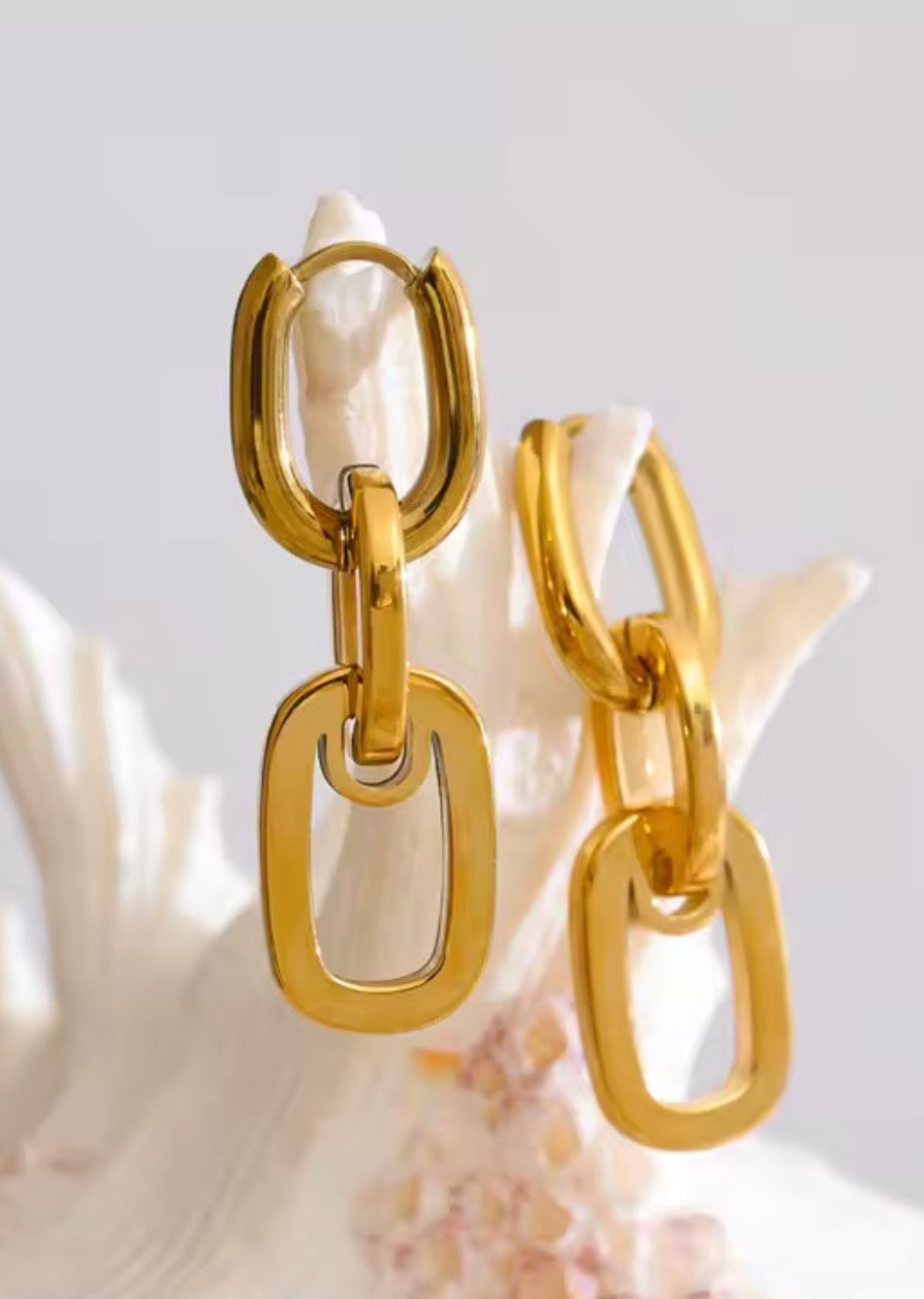 Gold-tone chain-link drop earrings with elongated oval links, displayed on a white seashell against a soft light gray background.