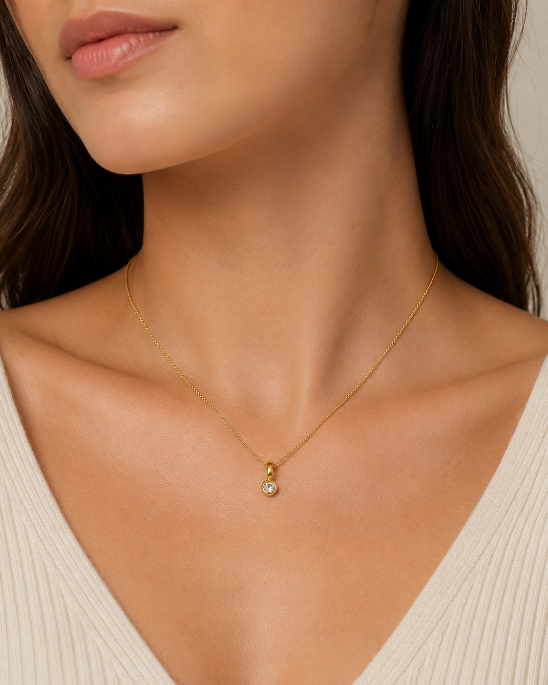 A close-up of a model wearing a delicate gold chain necklace with a small gold pendant featuring a clear round gemstone, paired with a cream-colored ribbed top.