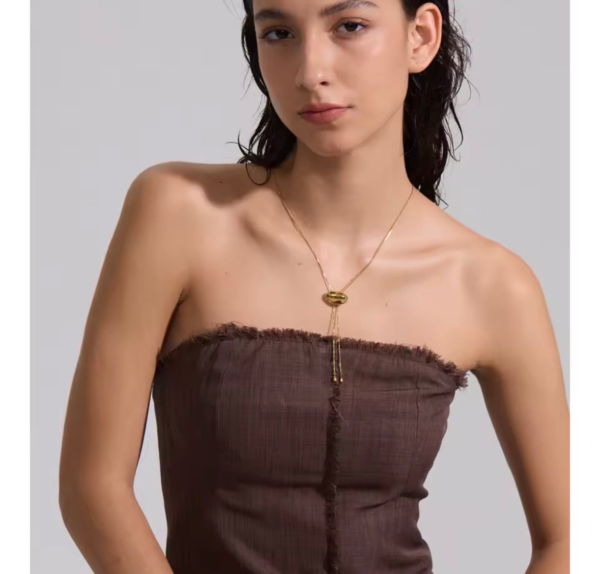 A woman wearing a dark brown strapless top and a gold lariat necklace with a textured oval pendant, photographed against a neutral gray background.