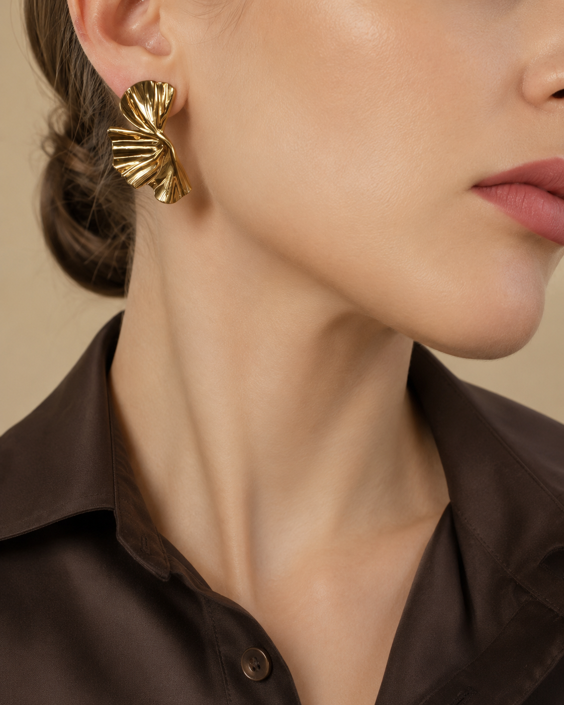 A close-up of a model wearing a textured gold earring with a sculptural, layered fan or leaf design. The model is wearing a dark brown satin button-up shirt against a solid beige background.