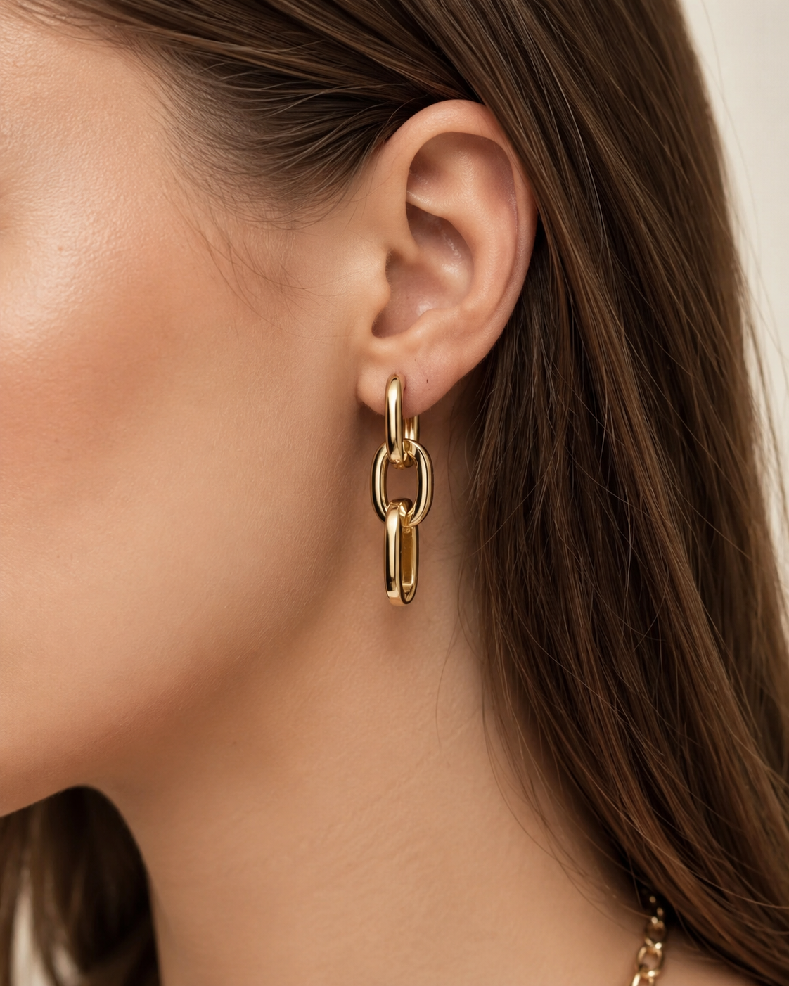 A close-up of a model's ear wearing a gold link drop earring, with her brown hair and neck visible against a soft light background.