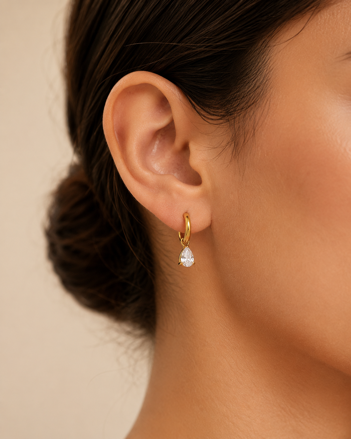 A close-up of a model's ear wearing a delicate gold huggie hoop earring with a small teardrop-shaped clear crystal drop.