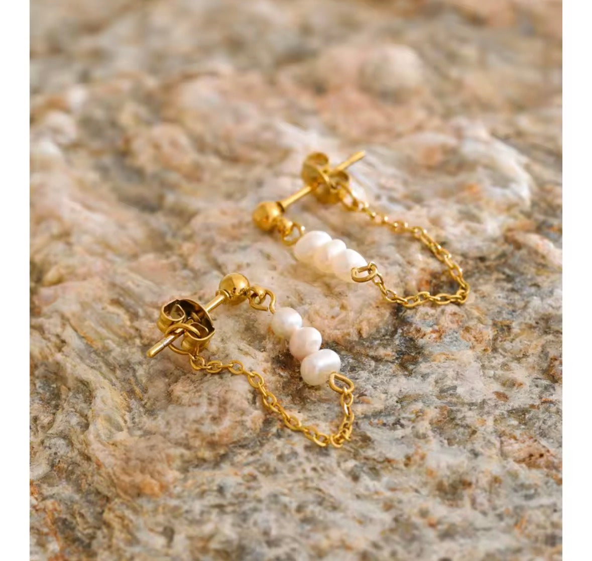 A pair of gold hoop earrings with small white pearl bead drops, displayed on a textured beige and brown stone surface.