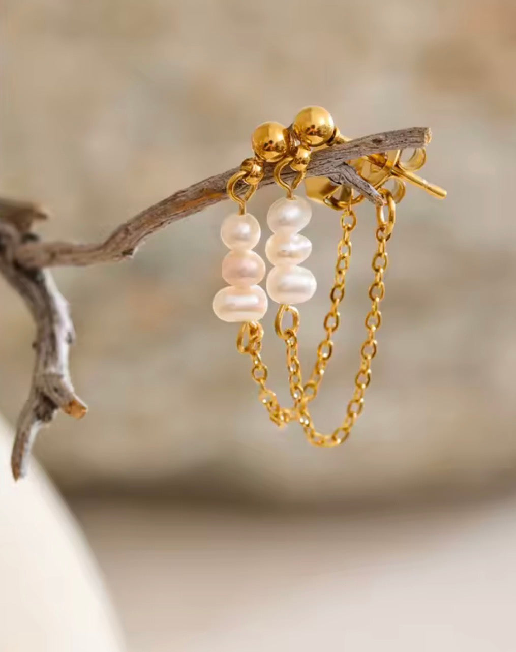 Gold dangle earrings with three white pearl beads and short gold chains, hanging from a small branch against a soft beige background.