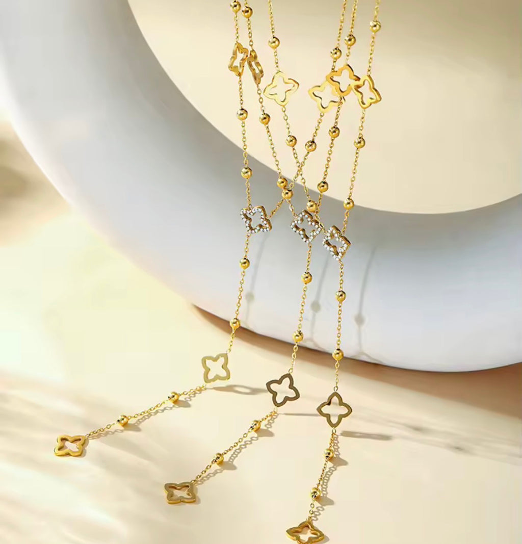 THE BLOOM NECKLACE A gold-toned multi-strand necklace with repeating clover-shaped charms, some outlined in gold and others set with small clear stones, displayed on a white curved jewelry stand.
