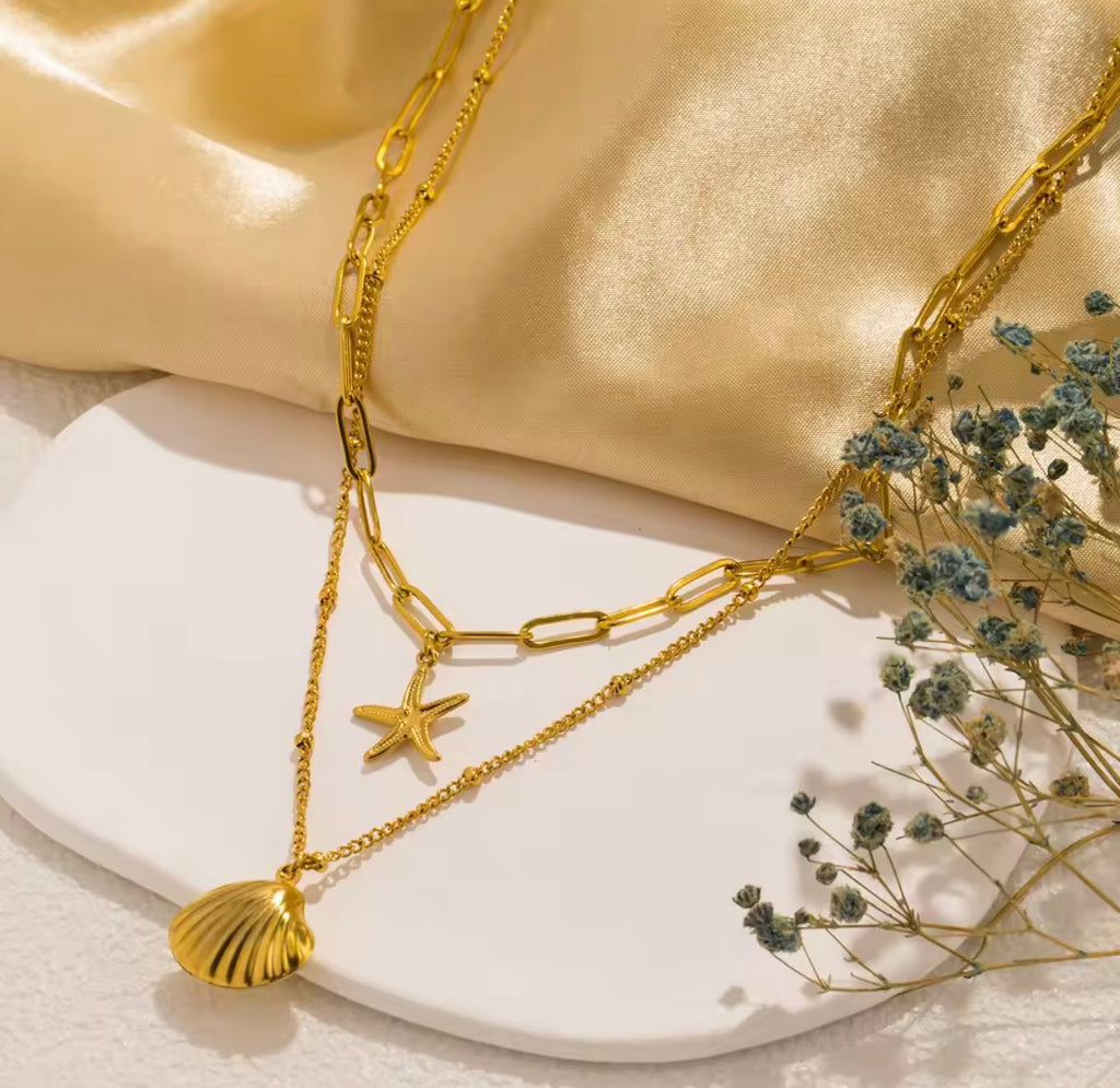The Calypso Necklace: Gold chain necklace with starfish and shell pendants on a beige fabric background