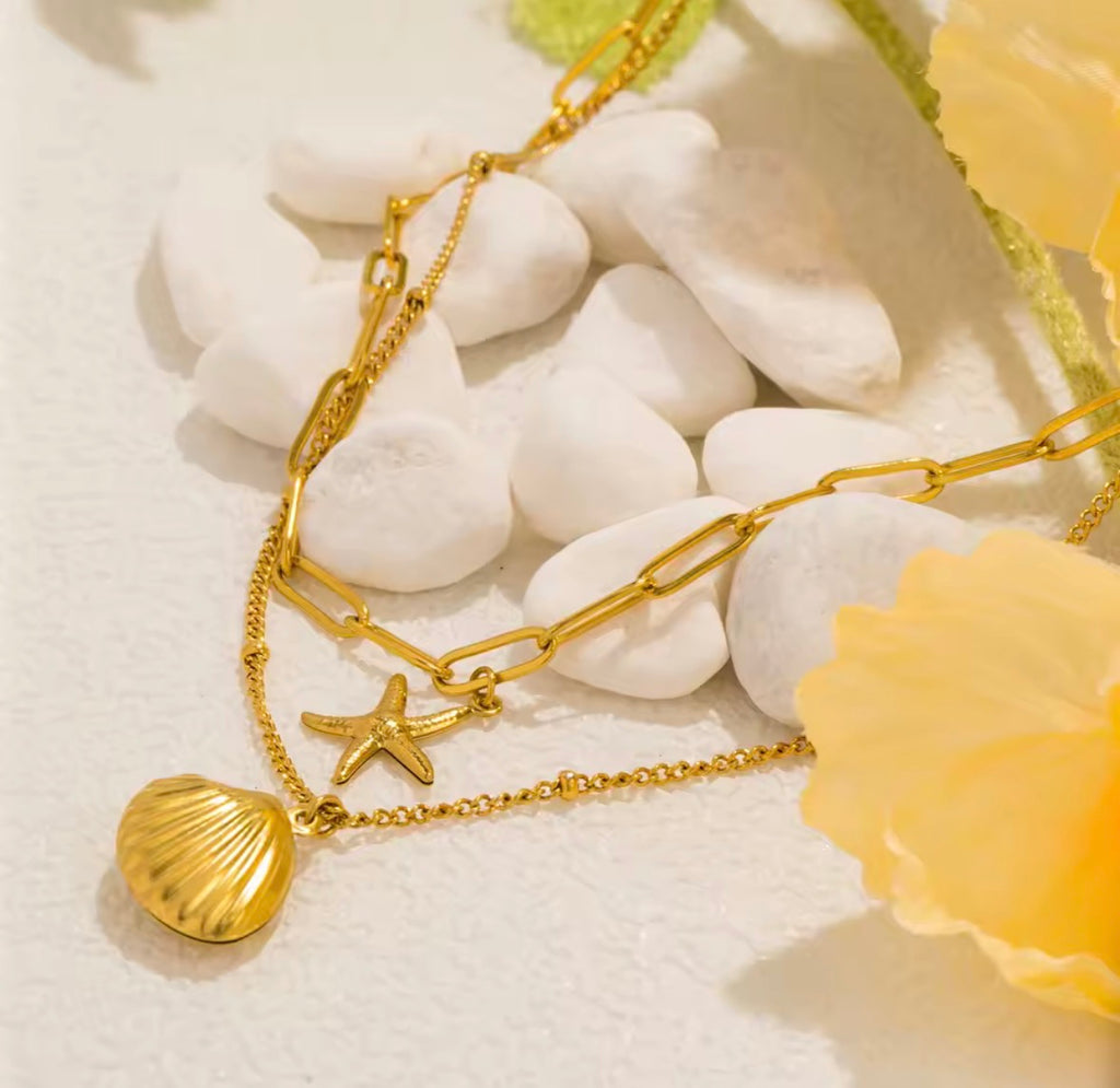 The Calypso Necklace: Gold layerd necklace with shell and starfish pendants on a white stone background