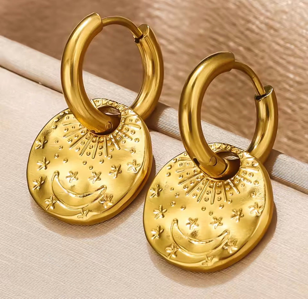 The Celeste Hoops Gold hoop earrings with moon and star designs on a beige surface