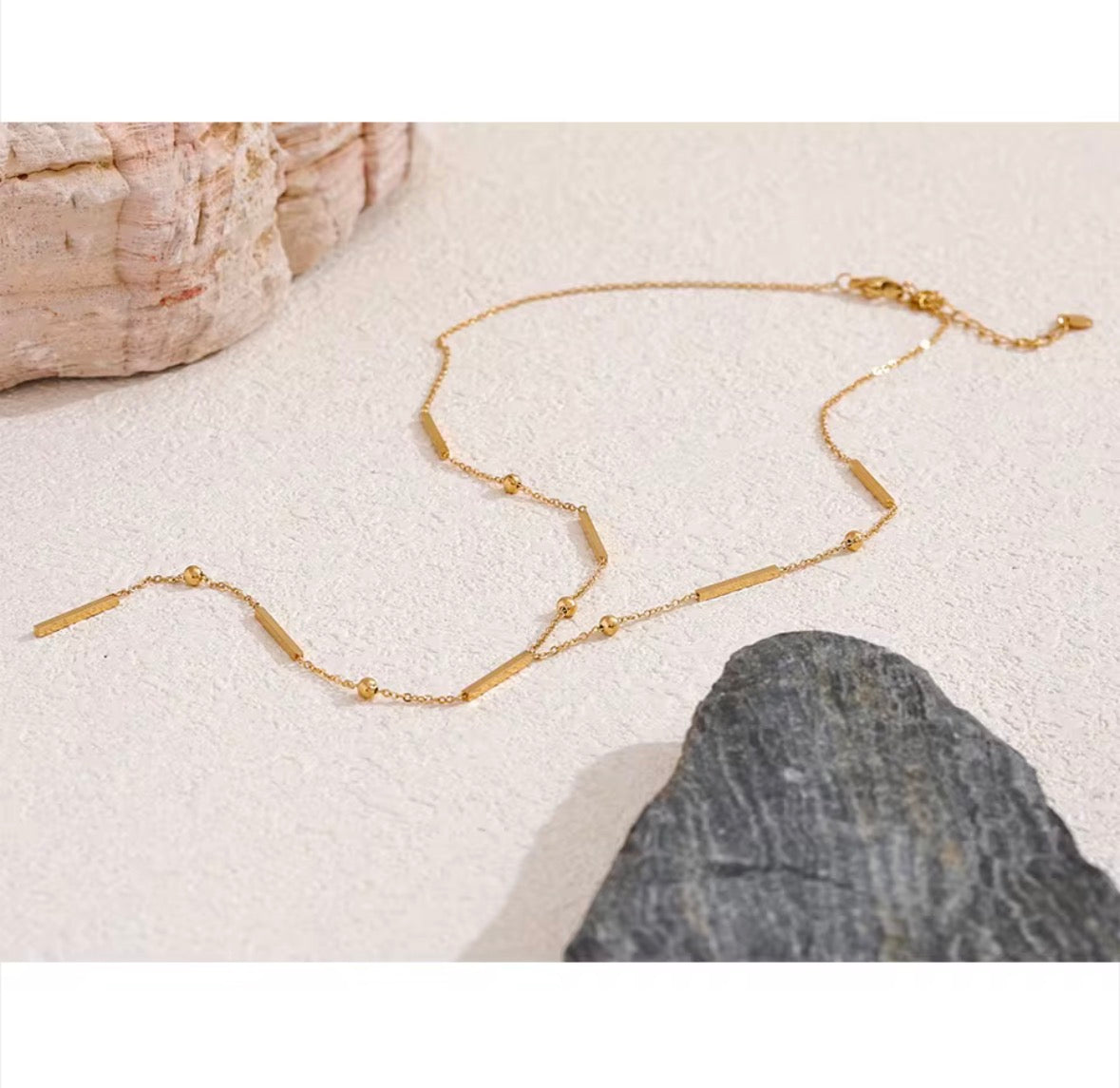 The Elisse Necklace A delicate gold chain necklace with alternating small round beads and polished rectangular bar links, displayed on a textured light beige surface with decorative stones.