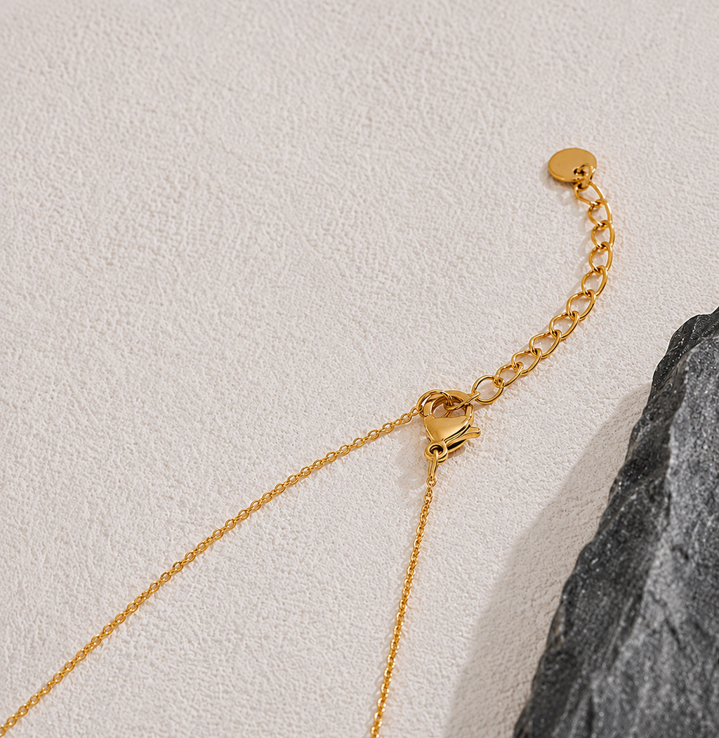 The Elisse Necklace Close-up of a gold chain necklace with a lobster clasp, extender chain, and round tag, laid on a textured off-white surface next to a dark gray stone.