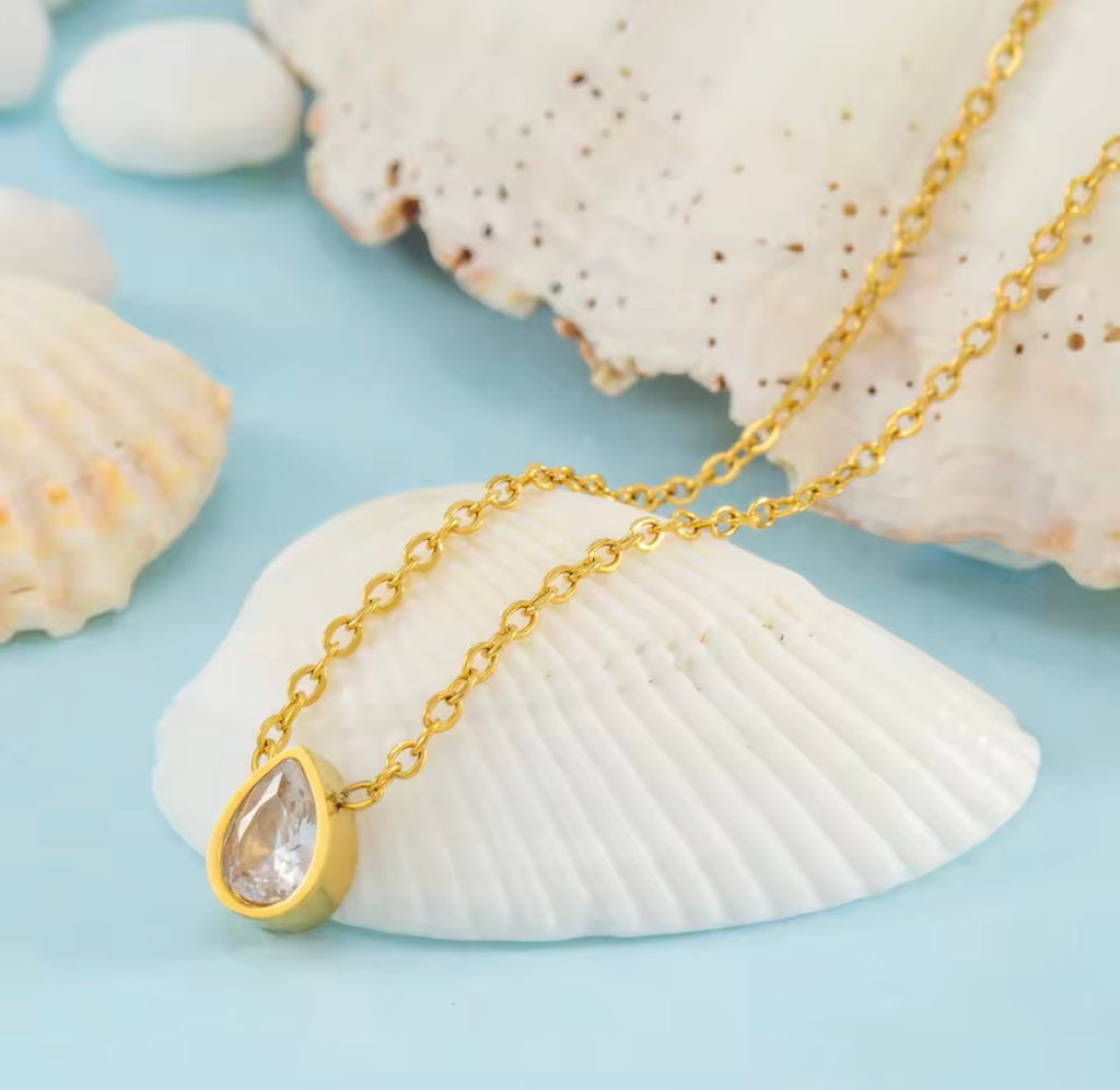 Gold necklace with a teardrop-shaped pendant on a light blue background with seashells.