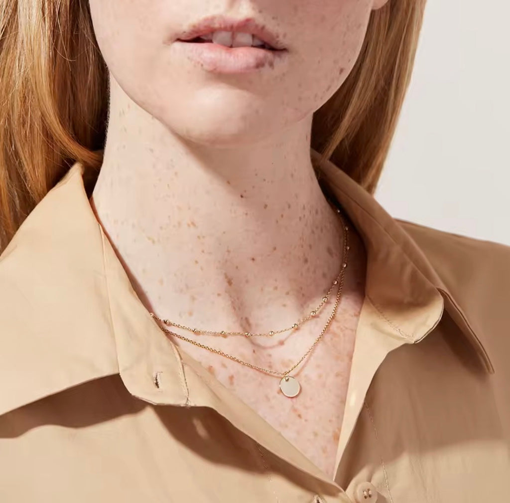 The Evienne Necklace  Close-up of a person wearing a beige shirt and gold necklaces with a neutral background