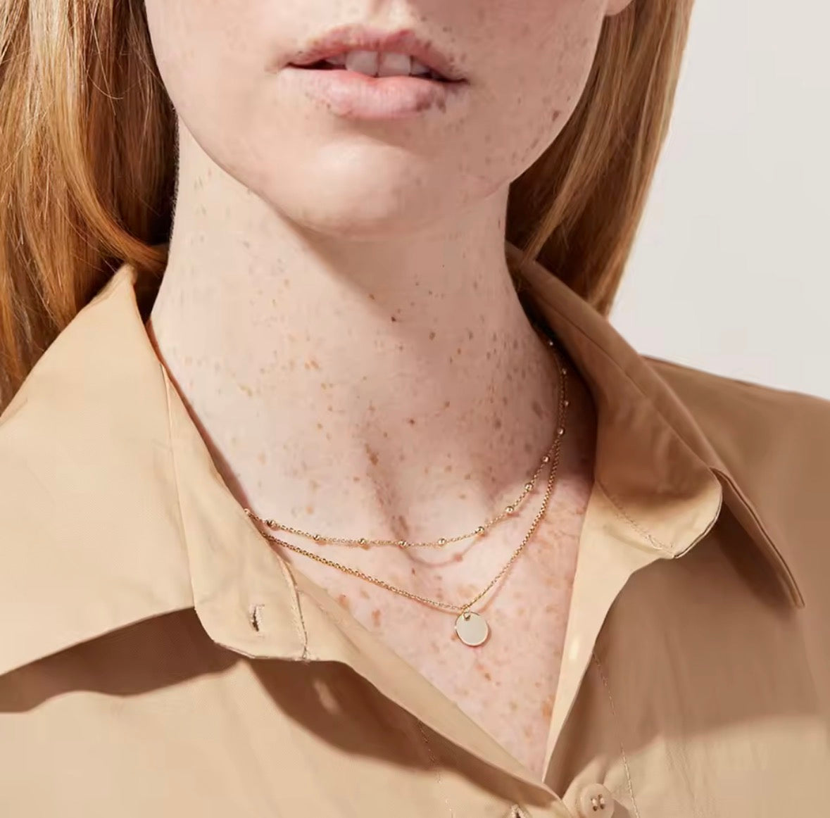 The Evienne Necklace  Close-up of a person wearing a beige shirt and gold necklaces with a neutral background