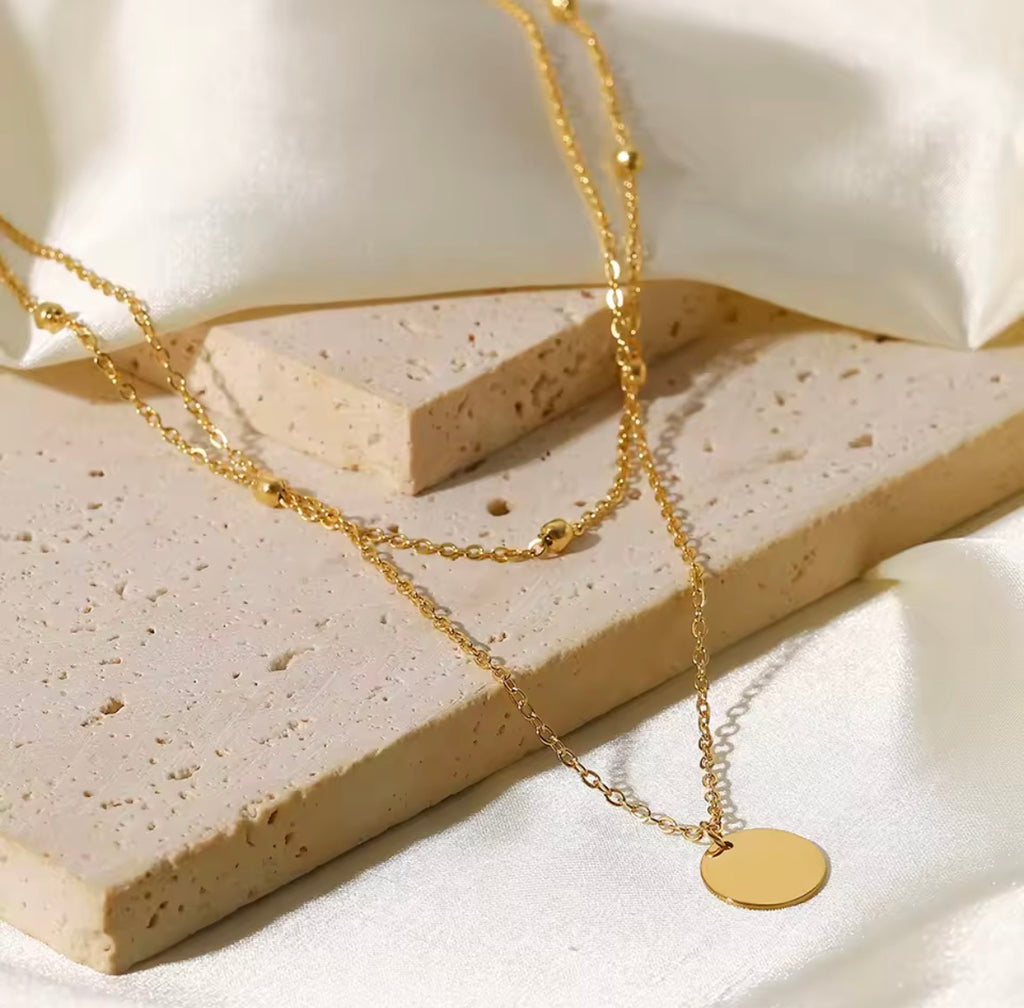 The Evienne Necklace  Gold necklace with a round pendant on a light stone surface