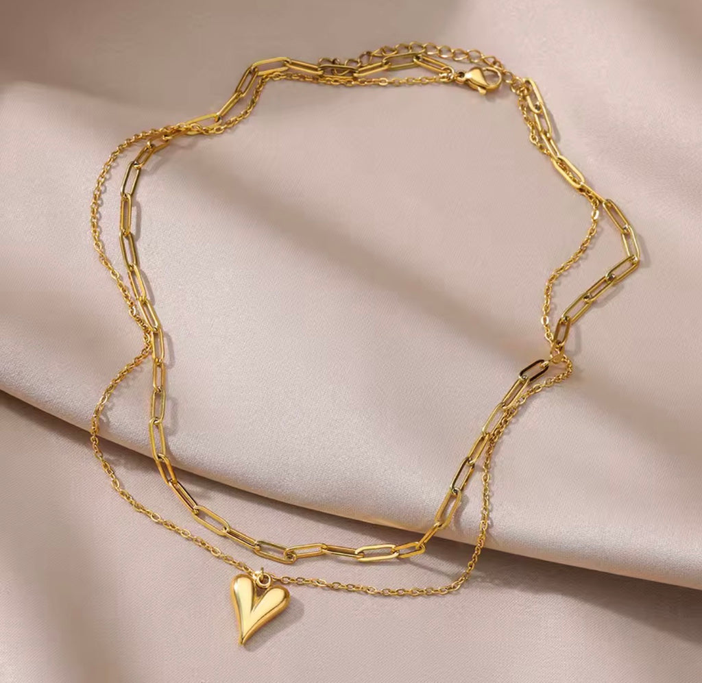 The Fiera Necklace, Gold chain bracelet with a heart pendant on a beige fabric background