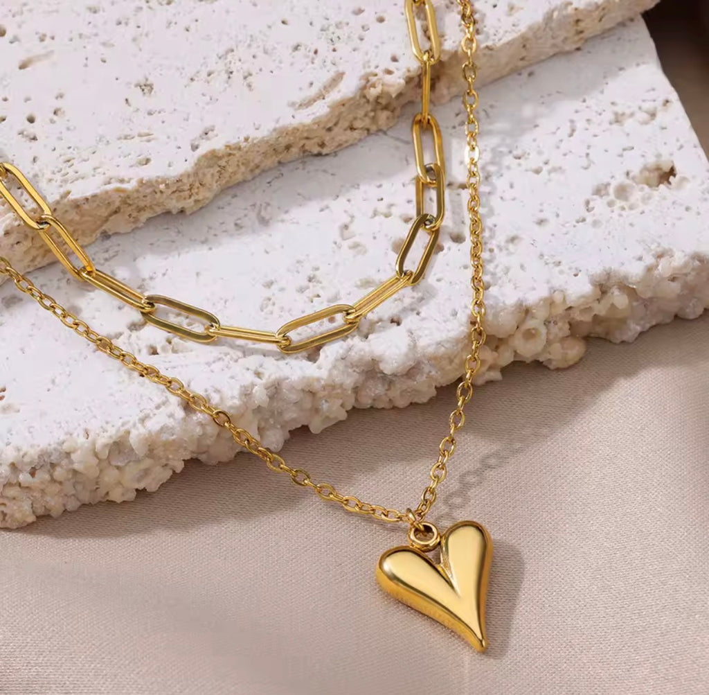 The Fiera Necklace Gold chain necklace with a heart pendant on a textured surface