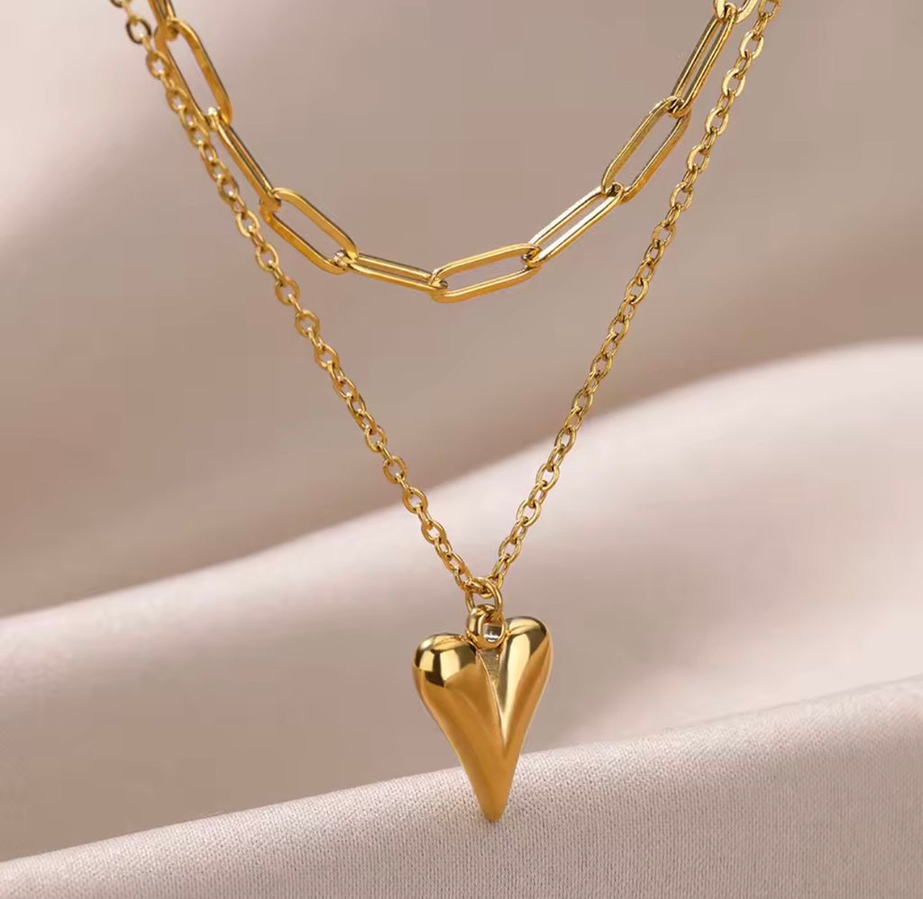 The Fiera Necklace Gold heart-shaped pendant necklace on a beige background