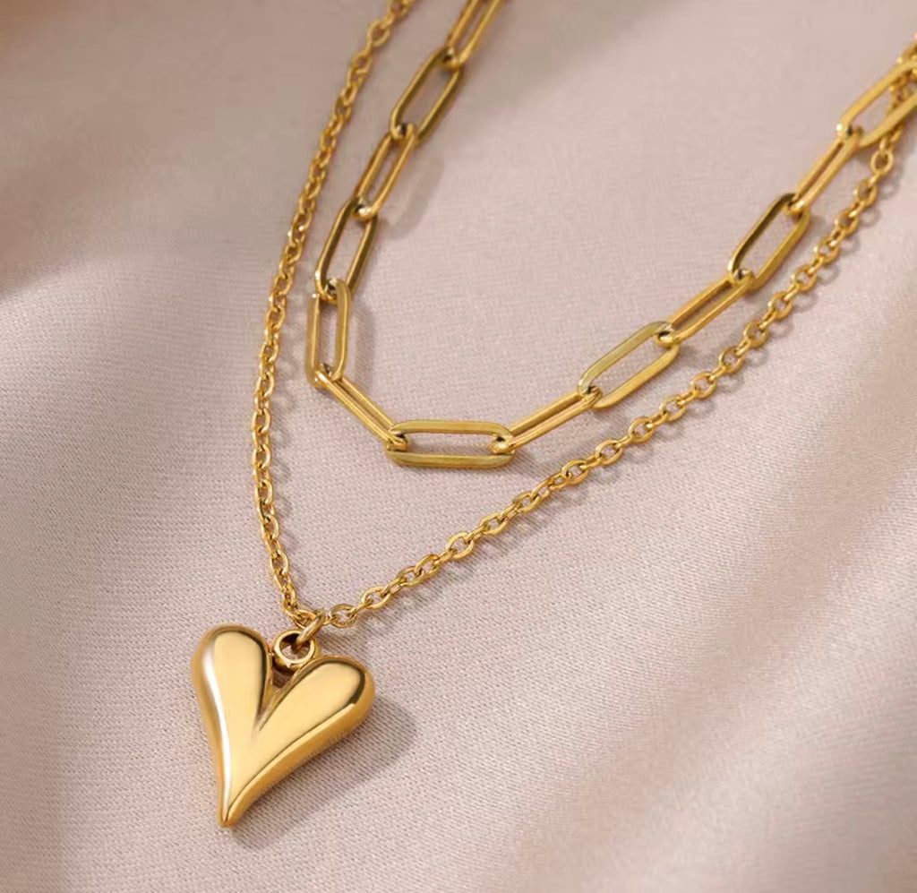 The Fiera Necklace, a gold heart-shaped pendant necklace on a beige fabric background