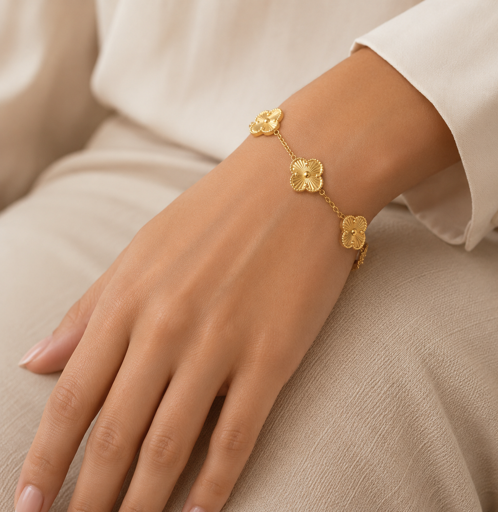 The Fiore Bracelet  A delicate gold chain bracelet with three textured flower charms worn on a woman's wrist, shown against a soft beige fabric background.