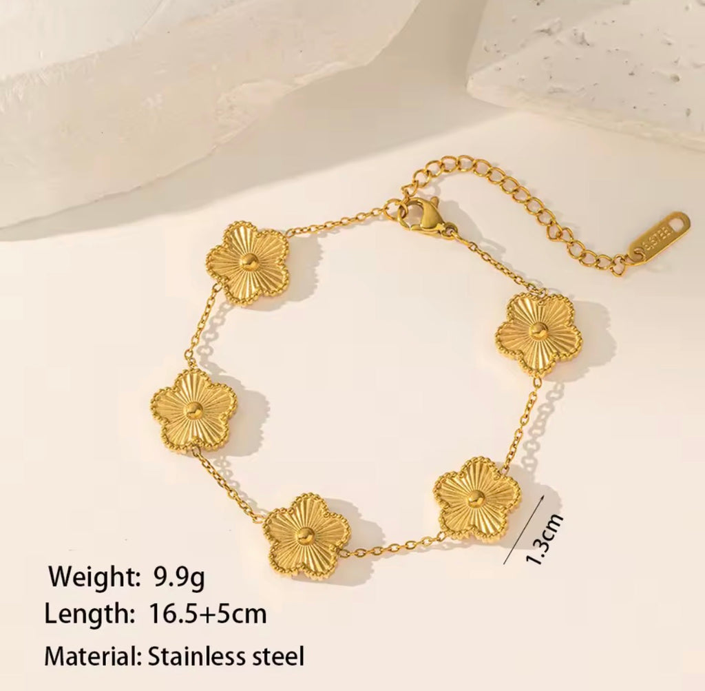 The Fiore Bracelet  A gold-tone stainless steel bracelet with five textured flower charms, shown on a light beige background with text detailing its weight, length, and material.