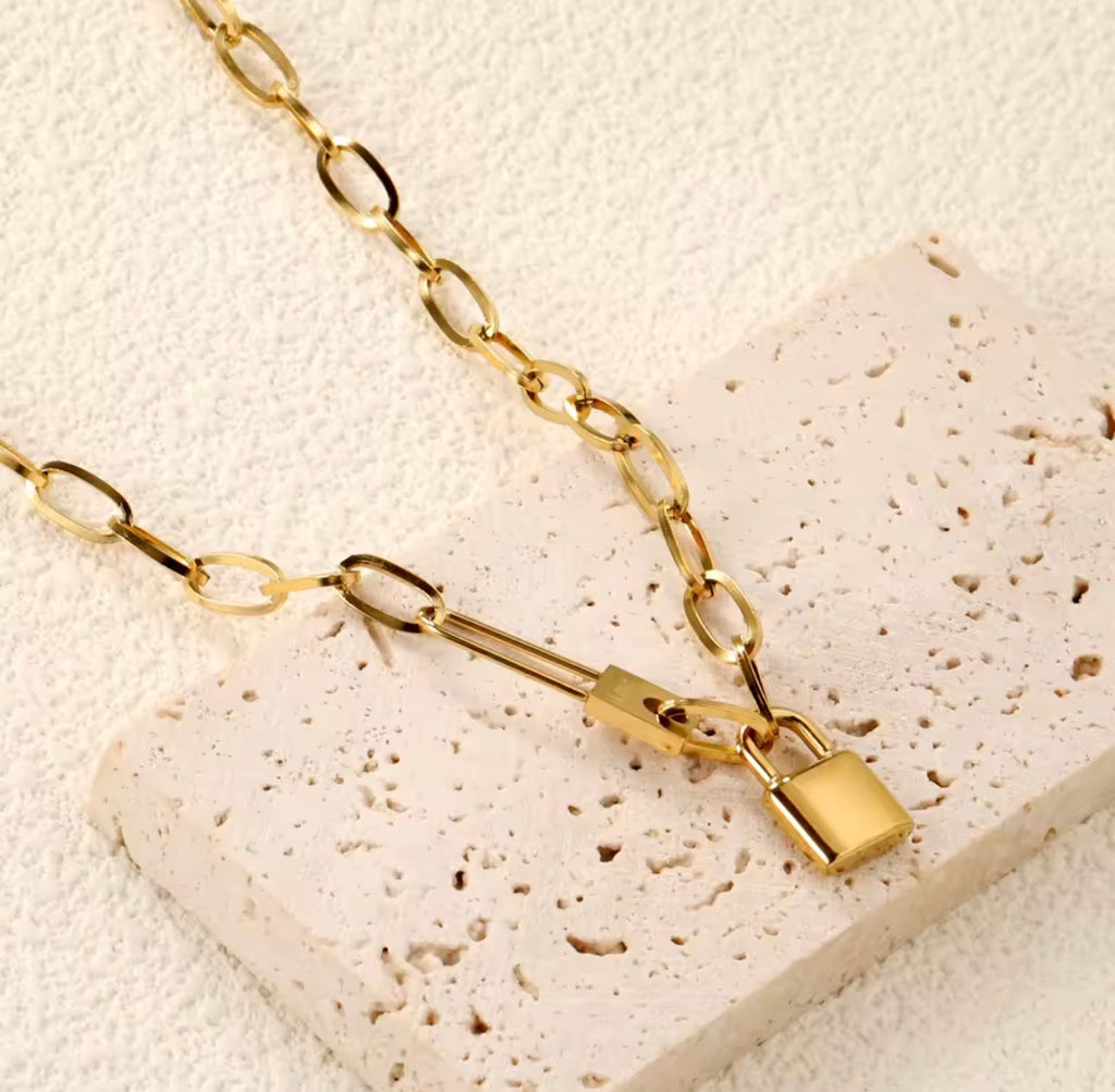 The Heartlock Necklace Gold chain necklace with a lock pendant on a textured beige surface