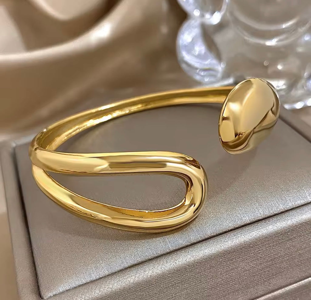 The Imperia Bangle Gold bracelet on a beige surface with a blurred background