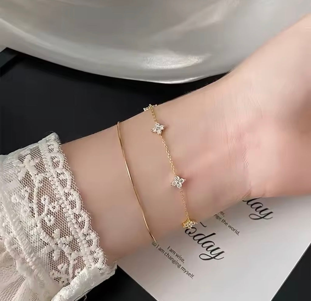 The Linnea bracelet  A close-up of a person's wrist wearing two delicate gold bracelets: one is a fine chain with small butterfly charms, and the other is a simple curved band. The person is wearing a white lace sleeve, and the background includes a white plate and a printed card.
