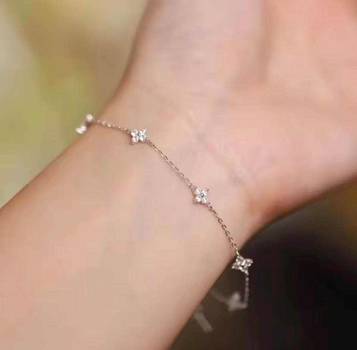 The Linnea bracelet  A delicate silver chain bracelet with three small flower charms, shown worn on a person's wrist against a soft, blurred background.