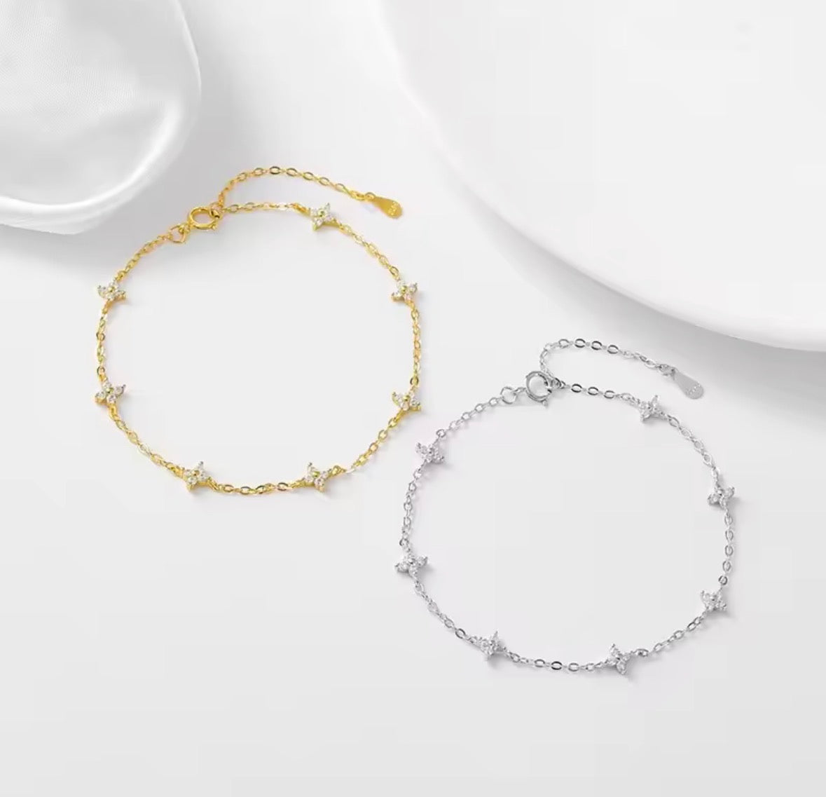 The Linnea bracelet Two delicate chain anklets with small star charms, one in gold and one in silver, displayed on a clean white surface with soft shadows.