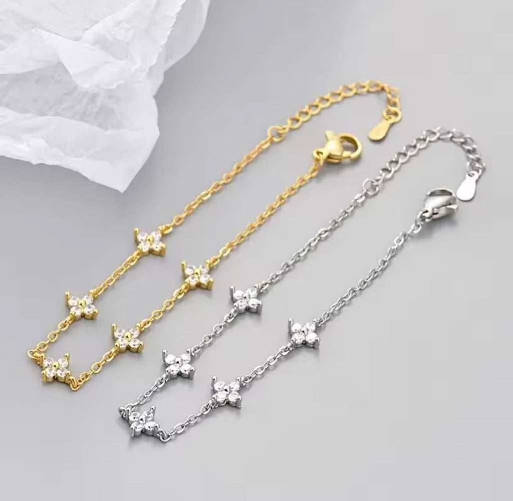 The Linnea bracelet Two delicate chain bracelets with small flower motifs, one in yellow gold and one in silver, displayed on a light gray surface with a white textured fabric in the corner.