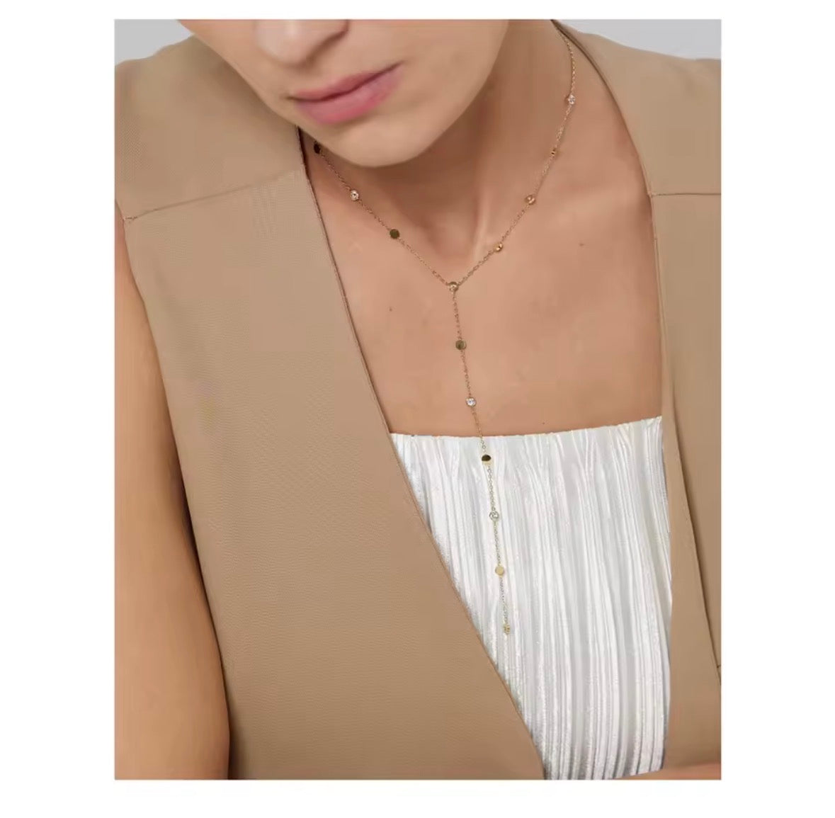The Orbit Necklace  A close-up of a model wearing a delicate gold Y-necklace with small gold and silver beads, styled over a white pleated top and a beige blazer.