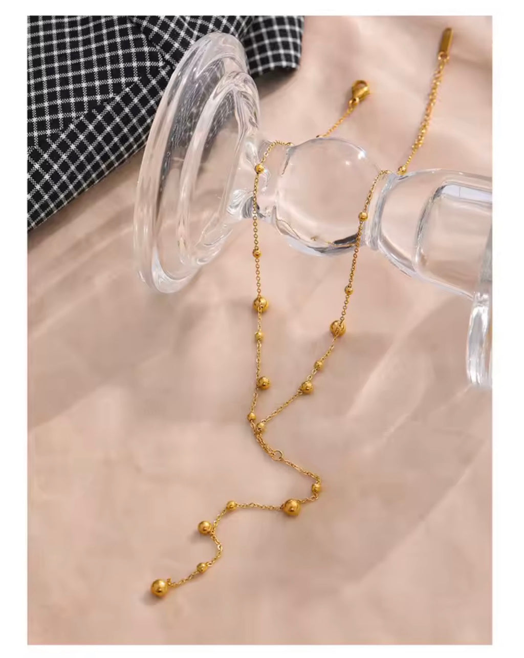 The Orbit Necklace A delicate gold beaded necklace draped over a clear glass stand on a soft beige fabric surface, with a black-and-white checkered fabric corner visible in the upper left.