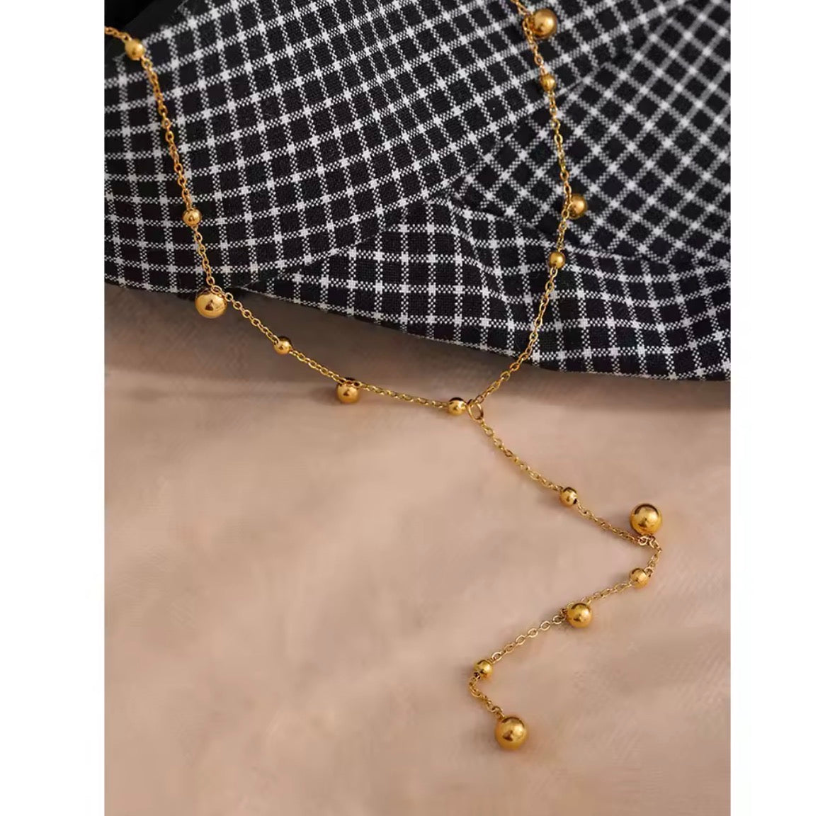 The Orbit Necklace  A gold Y-necklace with small beaded segments and larger polished ball charms, draped over a black and white checkered fabric and a beige surface.