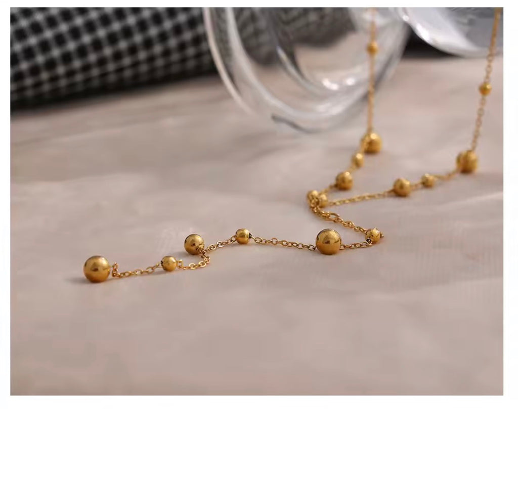 The Orbit Necklace  A gold beaded chain necklace with alternating small and larger spherical beads, displayed on a light beige fabric surface with a glass bowl and a black-and-white checkered fabric in the background.