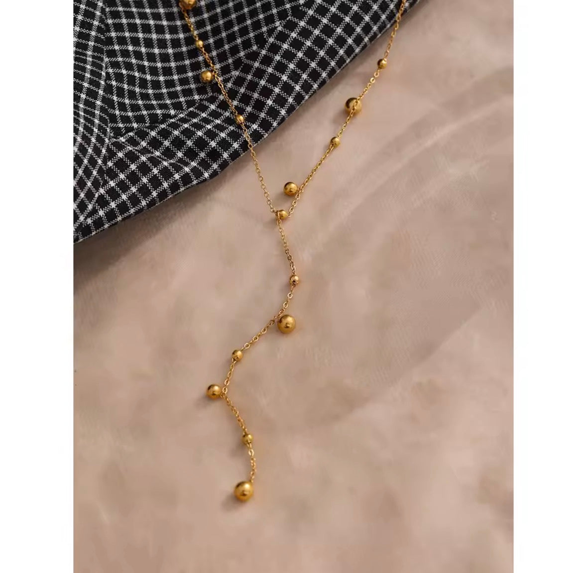 The Orbit Necklace A gold beaded necklace with a delicate chain and small spherical beads, styled on a soft beige fabric next to a black and white checkered fabric.