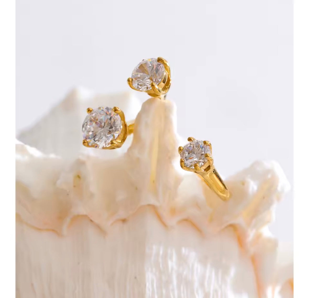 Three gold rings with round-cut clear gemstones displayed on a white seashell against a light gray background.