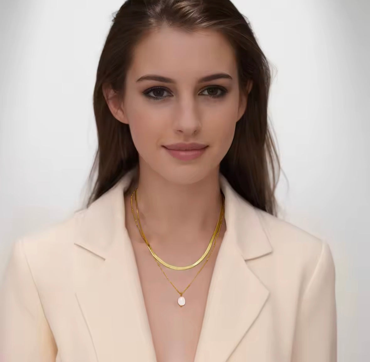 A woman wearing a beige blazer and gold necklaces against a white background. Woman Wearing The Aurelia Double Layer Gold Pearl Necklace